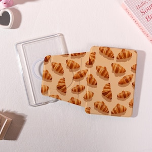 May include: A clear tablet case and a tan tablet cover with a pattern of brown croissants. The case is open, revealing the interior. The background is a light beige color.