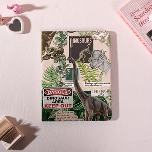 Może przedstawiać: Notatnik o tematyce dinozaurów. Okładka zawiera ilustracje dinozaurów, paproci i tekst "DINOSAURS". Zawiera znak "DANGER DINOSAUR AREA KEEP OUT" i tekst "LIFE FINDS A WAY."