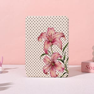 May include: A rectangular case with a cream-colored background covered in small black polka dots. Two pink lilies with green leaves are featured on the case. The lilies have a detailed watercolor design.