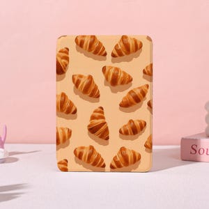 May include: A rectangular phone case featuring a repeating pattern of golden-brown croissants on a light tan background. The croissants are detailed with visible layers and textures, creating a realistic appearance. The case has rounded corners.