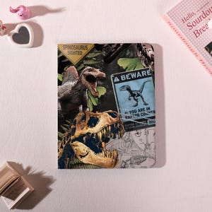 May include: A notebook with a dinosaur theme. The cover features a Spinosaurus, a dinosaur skull, and a sign that reads "BEWARE YOU ARE IN RAPTOR COUNTRY". The notebook also has the text "SPINOSAURUS SIGHTED".