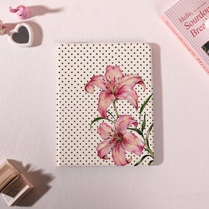 May include: A notebook with a white cover featuring a black polka dot pattern and a watercolor illustration of pink lilies. The lilies have green stems and leaves. The notebook is on a white surface.