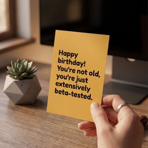 May include: A rectangular yellow greeting card with black text that reads: Happy birthday! You're not old, you're just extensively beta-tested. The card is held in a hand, positioned against a wooden desk surface with a small succulent plant.