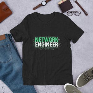May include: Dark grey t-shirt with the text "NETWORK ENGINEER" in green and white. The shirt also includes the phrase "I solve problems you don't know you have." The shirt is displayed on a light grey surface.