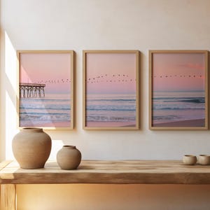 May include: Three framed beach prints with a pink and blue color palette. The prints depict a pier, ocean waves, and birds in flight. Two beige ceramic vases and three small bowls sit on a wooden table.