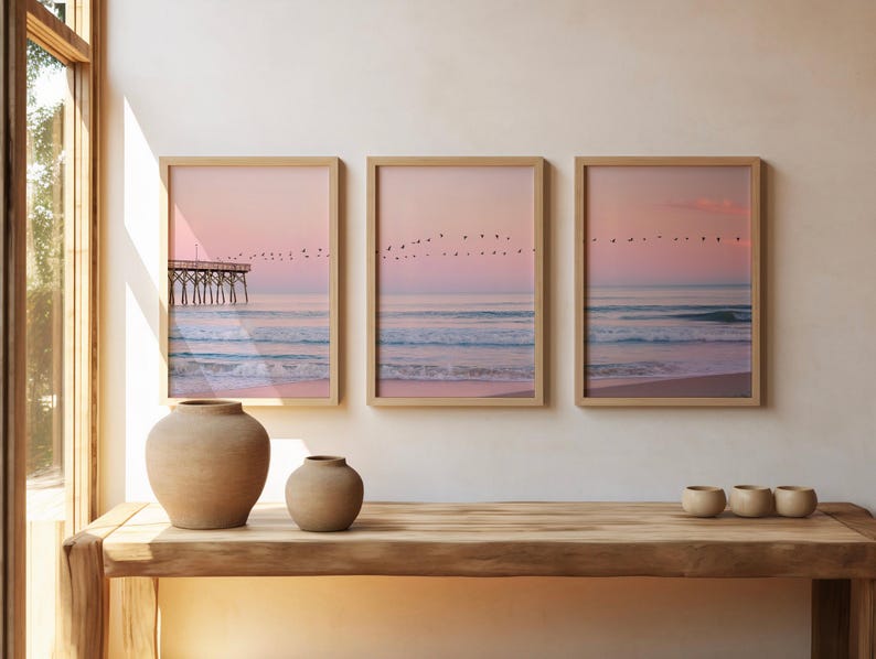 May include: Three framed beach prints with a pink and blue color palette. The prints depict a pier, ocean waves, and birds in flight. Two beige ceramic vases and three small bowls sit on a wooden table.