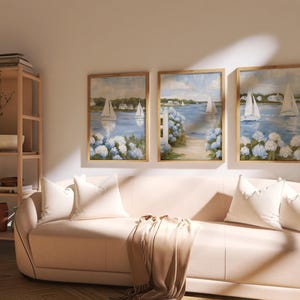 May include: Three framed landscape paintings with sailboats on a blue body of water, surrounded by white and blue flowers. The paintings are displayed above a light pink sofa with white pillows and a tan throw blanket. A wooden shelf is visible on the left.