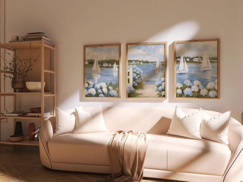 May include: Three framed landscape paintings with sailboats on a blue body of water, surrounded by white and blue flowers. The paintings are displayed above a light pink sofa with white pillows and a tan throw blanket. A wooden shelf is visible on the left.