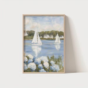 May include: A framed painting depicts two sailboats on a blue body of water, with white houses and green trees in the background. White flowers are in the foreground. The artwork has a light wood frame.