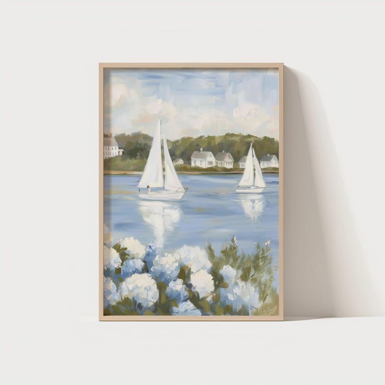 May include: A framed painting depicts two sailboats on a blue body of water, with white houses and green trees in the background. White flowers are in the foreground. The artwork has a light wood frame.