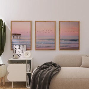 May include: Three framed art prints depicting a serene beach scene with a pier, birds, and a pastel pink and blue sky. A white nightstand with a clock and the text "KEEP IT CLASSY" is in front of the prints. A cactus and a beige sofa are also in the image.