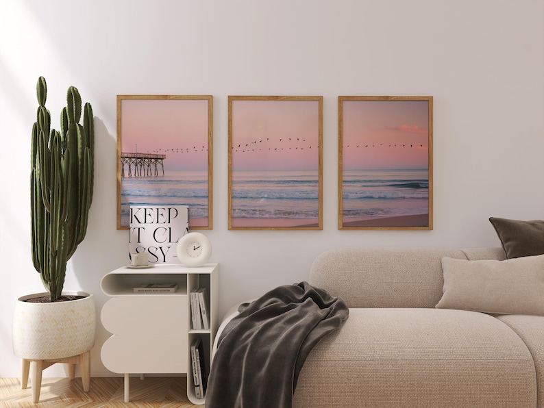 May include: Three framed art prints depicting a serene beach scene with a pier, birds, and a pastel pink and blue sky. A white nightstand with a clock and the text "KEEP IT CLASSY" is in front of the prints. A cactus and a beige sofa are also in the image.