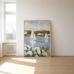 May include: A framed painting depicts a serene coastal scene with two sailboats on a blue body of water. White houses line the distant shore under a cloudy sky. White and blue flowers are in the foreground.