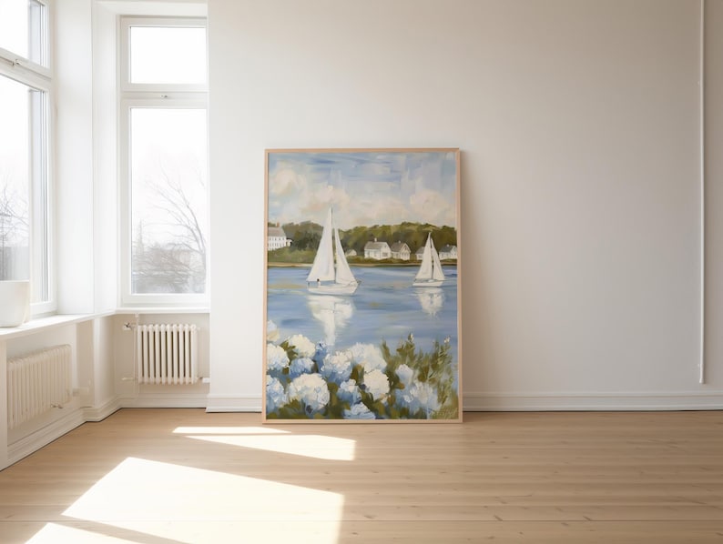 May include: A framed painting depicts a serene coastal scene with two sailboats on a blue body of water. White houses line the distant shore under a cloudy sky. White and blue flowers are in the foreground.