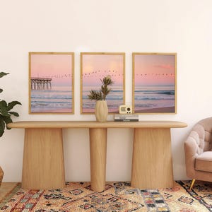 May include: Three framed beach scene prints with a pink and blue color palette hang above a wooden console table. A decorative vase, a small radio, and books sit on the table. A potted plant and a patterned rug complete the room's decor.