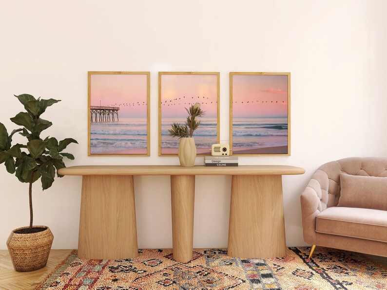 May include: Three framed beach scene prints with a pink and blue color palette hang above a wooden console table. A decorative vase, a small radio, and books sit on the table. A potted plant and a patterned rug complete the room's decor.