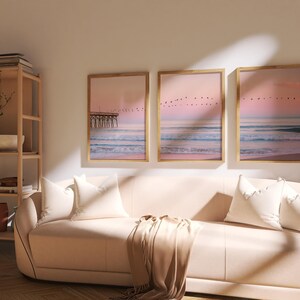 May include: Three framed landscape art prints depicting a beach scene with a pier, ocean waves, and a pink and purple sunset. The prints are displayed above a light pink sofa with white pillows and a tan throw blanket.