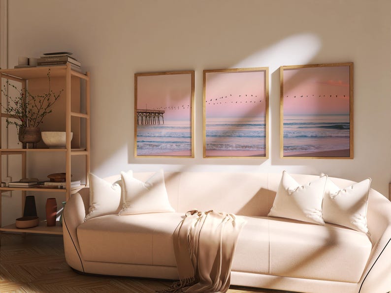 May include: Three framed landscape art prints depicting a beach scene with a pier, ocean waves, and a pink and purple sunset. The prints are displayed above a light pink sofa with white pillows and a tan throw blanket.