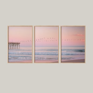 May include: Set of three framed prints depicting a serene beach scene at dawn or dusk. The sky transitions from pink to orange, with a flock of birds in flight. The ocean waves gently roll onto the sandy shore, and a pier is visible in one print.