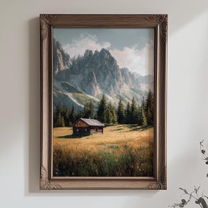 Könnte beinhalten: Gerahmtes Landschaftsgemälde mit einer Bergkette und einer kleinen Holzhütte in einem Feld. Das Kunstwerk hat einen braunen Holzrahmen mit dekorativen Eckdetails. Der Himmel ist eine Mischung aus Blau und Weiß.