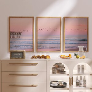 May include: Three framed beach scene prints with a pier, birds, and ocean waves. The prints are displayed above a white cabinet with gold hardware. A small speaker, snacks, fruit, and a pitcher are on the cabinet.