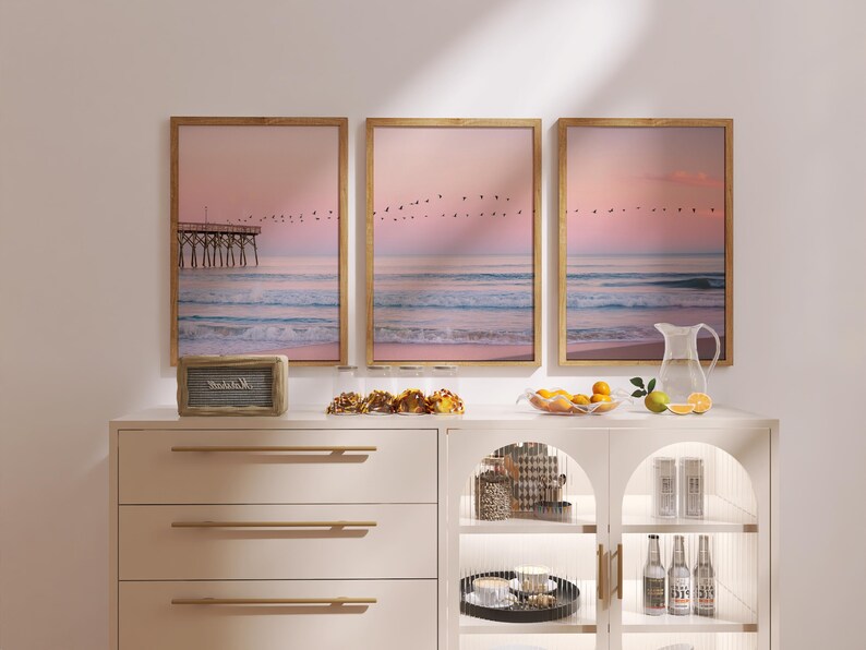 May include: Three framed beach scene prints with a pier, birds, and ocean waves. The prints are displayed above a white cabinet with gold hardware. A small speaker, snacks, fruit, and a pitcher are on the cabinet.