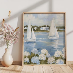 May include: A framed painting depicts two sailboats on a blue body of water, with white sails and a coastal landscape in the background. White and blue hydrangeas are in the foreground. The frame is made of light-colored wood.