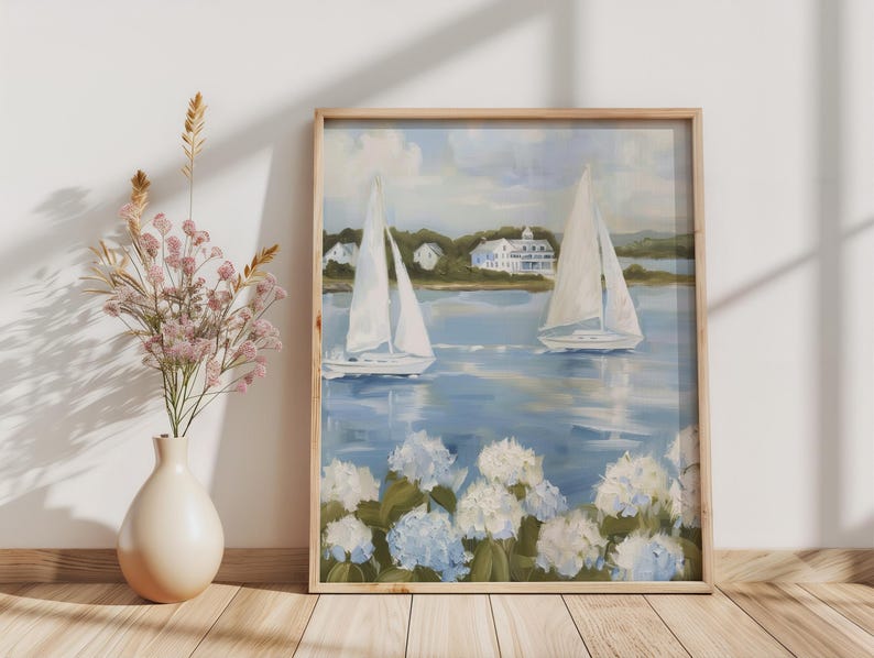 May include: A framed painting depicts two sailboats on a blue body of water, with white sails and a coastal landscape in the background. White and blue hydrangeas are in the foreground. The frame is made of light-colored wood.