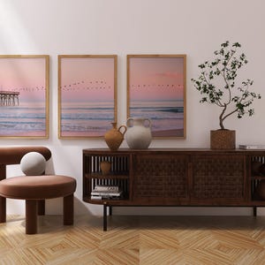 May include: A modern interior scene featuring three framed landscape prints with a pier and birds against a pink sky. A brown wooden cabinet, a chair, and decorative vases complete the decor. A potted plant adds a touch of nature.
