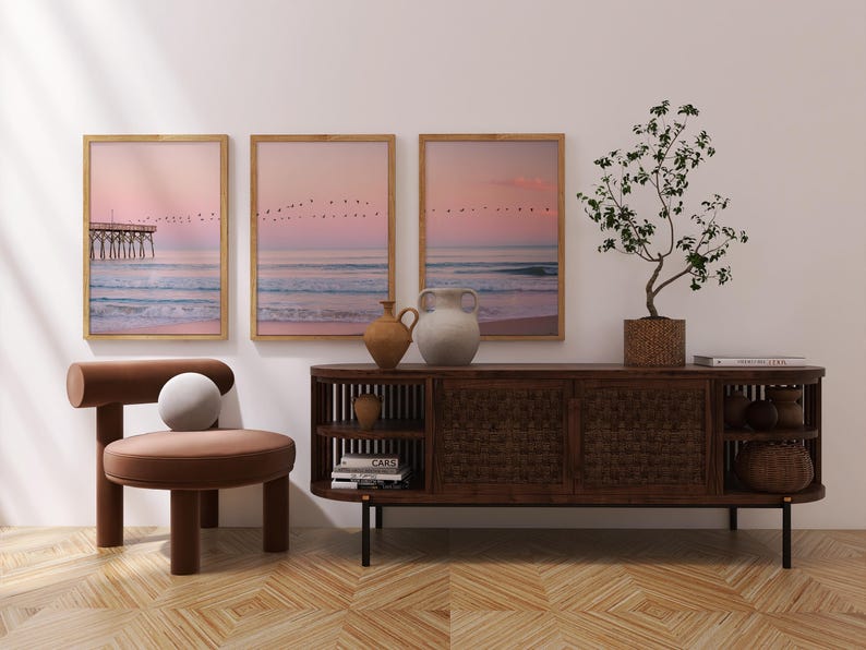 May include: A modern interior scene featuring three framed landscape prints with a pier and birds against a pink sky. A brown wooden cabinet, a chair, and decorative vases complete the decor. A potted plant adds a touch of nature.