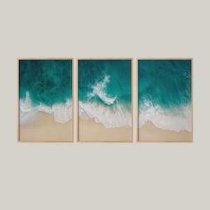 May include: Three framed wall art pieces featuring aerial views of a beach. Each piece shows turquoise ocean water meeting a sandy shore, with white waves breaking along the coast. The frames are a light brown color.