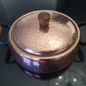 May include: A hammered copper pot with a wooden lid and handles. The pot has a shiny copper finish and a slightly rounded shape.