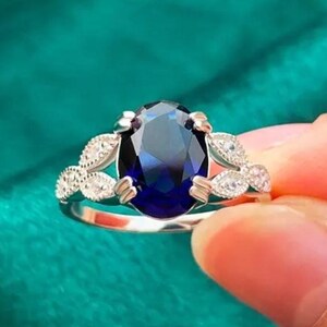 May include: A silver ring with a large, oval, deep blue gemstone. The ring has a delicate design with small, clear stones on either side of the main stone, set against a teal background.