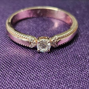 May include: A gold-coloured ring featuring a central, clear gemstone. The ring has a decorative band with small, clear accent stones on either side of the central stone. The ring is set against a purple fabric background.