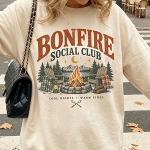 May include: A sand-colored crewneck sweatshirt featuring a graphic print with the text "BONFIRE SOCIAL CLUB" above an illustration of a campfire, two wooden chairs, pine trees, and mountains, with the text "COOL NIGHTS WARM FIRES" below.
