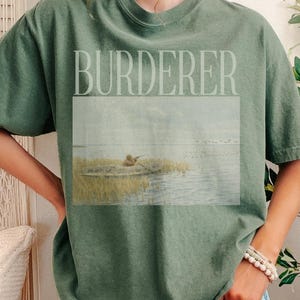 May include: An olive green t-shirt with the word "BURDERER" printed across the top. Below the text is a rectangular image of a person in a boat on a lake. The person is wearing a hat and is fishing. The shirt is a relaxed fit.