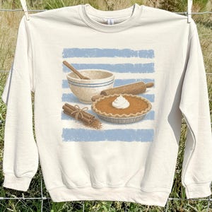 May include: A sand-colored crewneck sweatshirt featuring a central graphic print of a pumpkin pie with whipped cream, a wooden rolling pin, a mixing bowl, and cinnamon sticks, all set against a background of horizontal light blue distressed stripes.