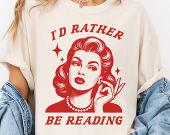 Op de afbeelding: Ivoor T-shirt met een rode afbeelding van een vrouw en de tekst "I'd Rather Be Reading". De afbeelding toont een vintage illustratie van een vrouw met rood haar, een parelketting en een handgebaar. Het shirt is een casual stijl.