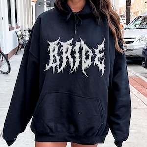 May include: Black hooded sweatshirt with the word "BRIDE" in a spiky, gothic font across the chest. The sweatshirt has a front pocket and a drawstring hood. The image is taken outdoors on a sidewalk.