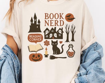 May include: Ivory-colored t-shirt featuring a Halloween-themed graphic. The design includes a haunted house, a black cat, a pumpkin, and the words "BOOK NERD" and "READING CORNER". The shirt is made of a soft material.
