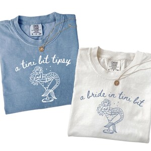 May include: Two t-shirts, one light blue and one off-white, with matching gold necklaces. The light blue shirt says "a tini bit tipsy" with a mermaid in a cocktail glass. The off-white shirt says "a bride in tini bit" with a similar design.
