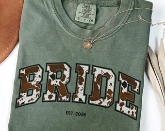 Può includere: Una maglietta verde oliva con la scritta "BRIDE" in un motivo marrone e bianco a stampa di mucca. Il testo "EST. 2026" è sotto la parola "BRIDE". Una collana d'oro con un ciondolo è sulla maglietta.