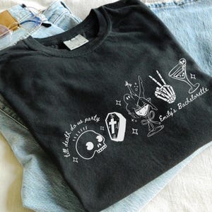 Może przedstawiać: Czarny t-shirt z białym nadrukiem przedstawiającym czaszkę, trumnę, kieliszek do martini, szkieletową dłoń oraz napisy "till death do us party" i "Emily's Bachelorette". Koszulka jest złożona na niebieskich dżinsach.