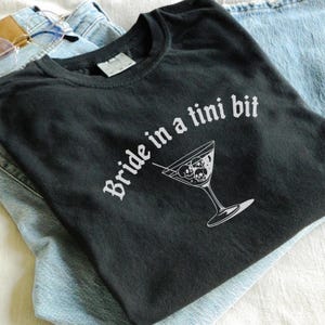 May include: Black t-shirt with the phrase "Bride in a tini bit" in white script. A martini glass graphic with skull-shaped olives is below the text. The shirt is folded on top of blue jeans.