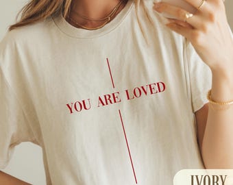 May include: Ivory-colored t-shirt with the text "YOU ARE LOVED" in red. The design includes a vertical line above and below the text. The word "IVORY" is displayed in a rectangular box at the bottom right.