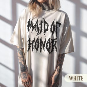 May include: Cream-colored t-shirt with the text "MAID OF HONOR" in a black, gothic font. The shirt has short sleeves and a relaxed fit. The word "WHITE" is displayed in a white box at the bottom right.