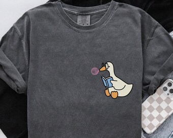 Puede incluir: Camiseta de manga larga gris oscuro con un dibujo de un ganso de dibujos animados con gafas y leyendo un libro mientras sopla una burbuja. El ganso es blanco con patas naranjas y un libro azul.