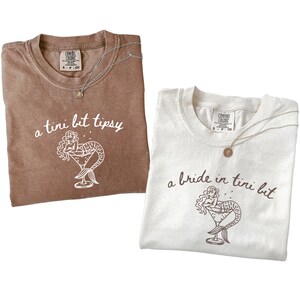 May include: Two t-shirts, one brown and one white, with the text "a tini bit tipsy" and "a bride in tini bit" printed on them. Each shirt features a mermaid illustration in a martini glass. Both shirts have a silver necklace.