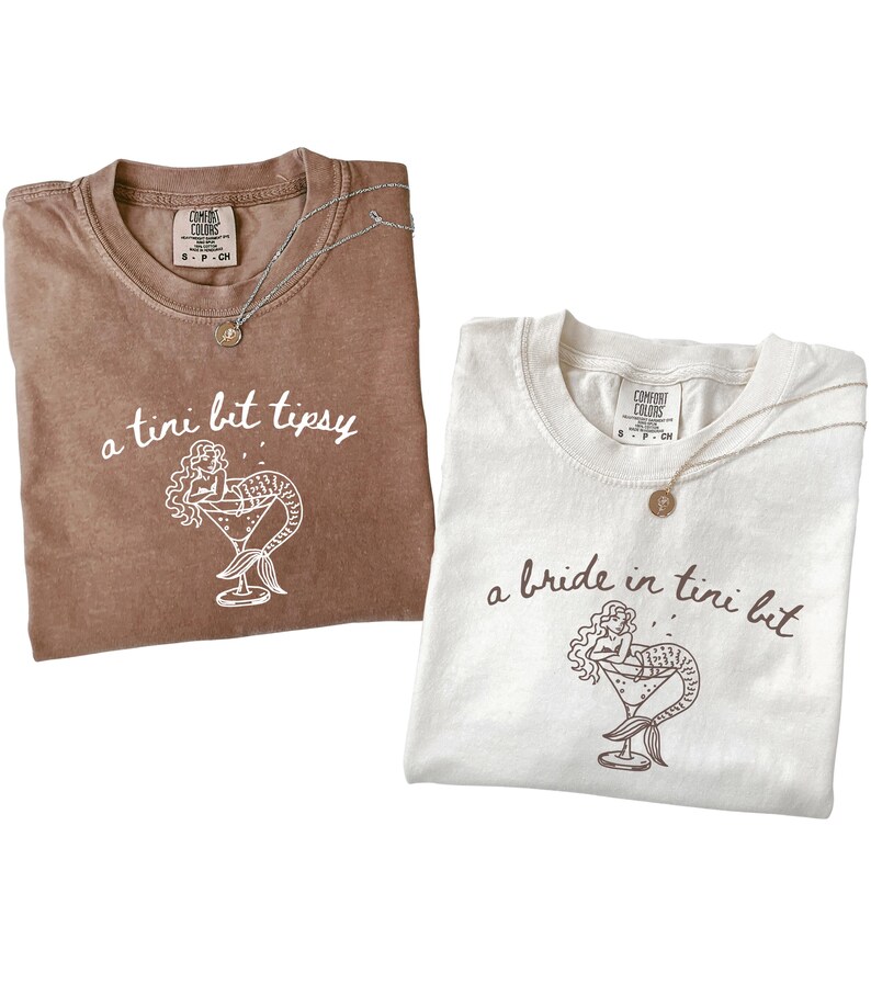 May include: Two t-shirts, one brown and one white, with the text "a tini bit tipsy" and "a bride in tini bit" printed on them. Each shirt features a mermaid illustration in a martini glass. Both shirts have a silver necklace.