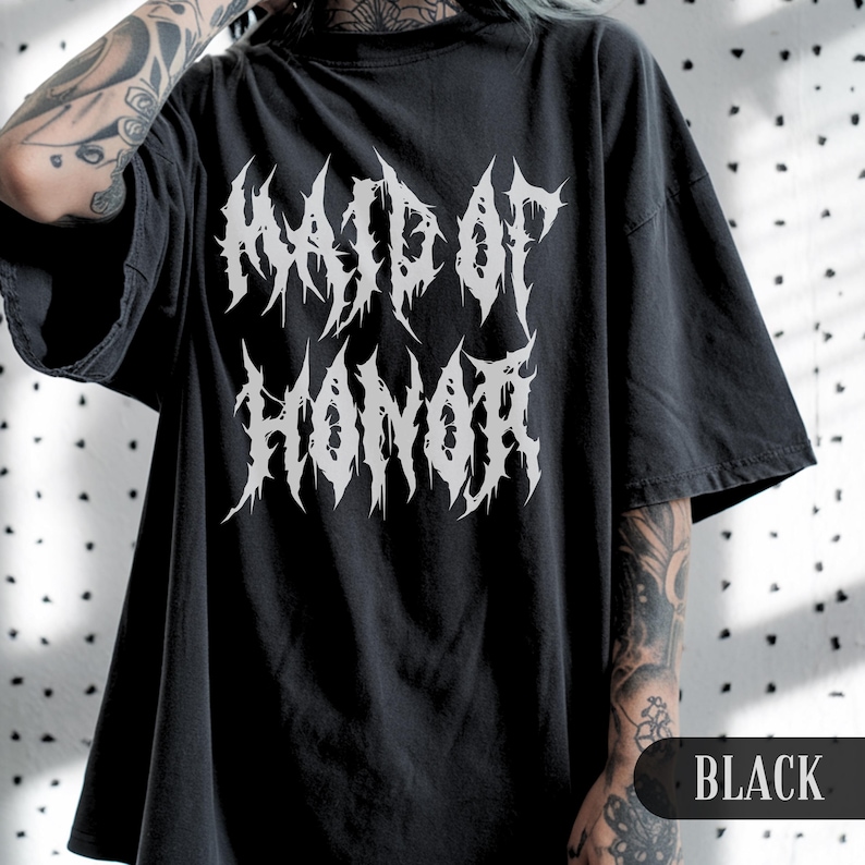 May include: Black oversized t-shirt with the text "MAID OF HONOR" in a white, gothic font. The shirt has short sleeves and a crew neck. The word "BLACK" is printed at the bottom right corner of the image.